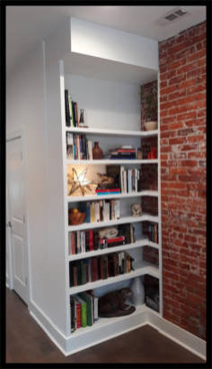 Custom white Maple bookcase nestled in a red brick nook with fixed-height shelving and high baseboard moldings.